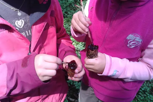 Zwei Kinder halten aufgebrochene Tannenzapfen, in denen sie rote Farbe aus zerdrückten Heidelbeeren rühren